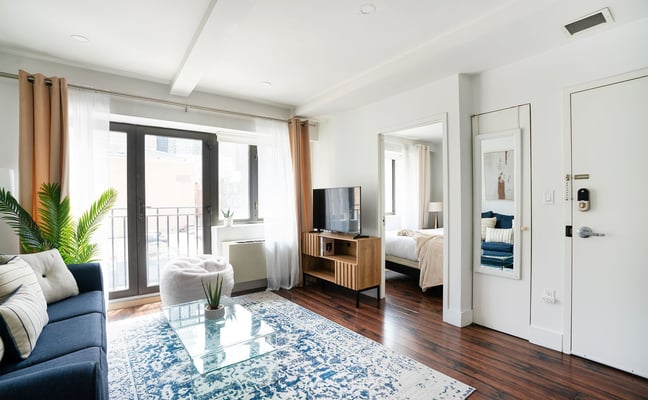 Bright furnished NYC apartment living room with blue sofa, glass coffee table, and balcony access